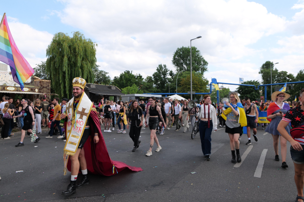 Eine Gruppe von Menschen marschiert bei der Gay Pride Parade 2018 mit einer Regenbogenflagge und Musikinstrumenten, umgeben von Laternenmasten, Bäumen, Hütten und einem bewölkten Himmel.