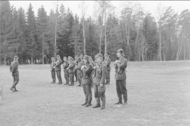 Schwarzes Bild einer Gruppe von Männern in militärischer Kleidung, die in einem Feld stehen und Gewehre halten, mit Bäumen und einem klaren Himmel im Hintergrund.