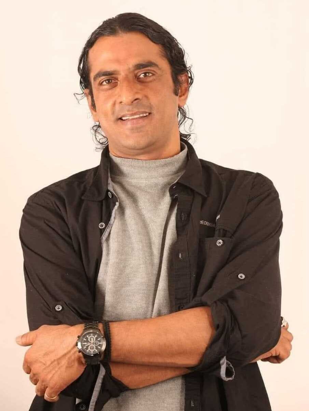 Ein Mann in einem schwarzen und aschfarbenen Kleid mit einer Uhr steht mit verschränkten Armen tief in Gedanken vor einem weißen Hintergrund.