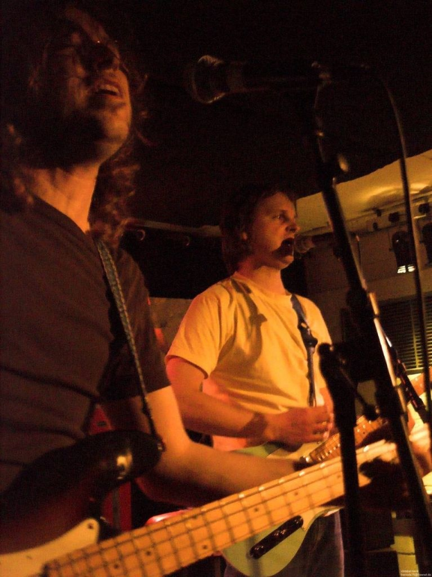 Ein Musikkonzert mit zwei Sängern; einer in einem weißen Hemd, der eine grüne Gitarre hält, und der andere in einem schwarzen Hemd mit Brille, der eine schwarze Gitarre hält, beide mit Mikrofonen, vor einem Hintergrund aus Scheinwerfern und anderen Instrumenten.