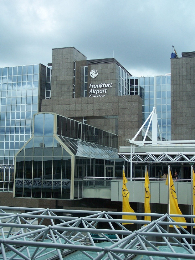 Frankfurt Airport in Frankfurt, Germany, featuring a large glass building with text, surrounded by yellow flags and iron rods, under a sky with white clouds.