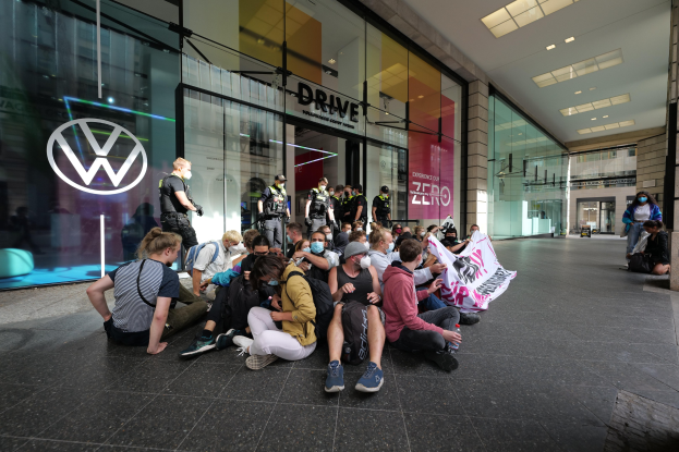 Eine Gruppe von Menschen sitzt vor einer Glasfront eines Volkswagen-Händlers auf dem Boden und hält eine Fahne mit Text, unter Deckenleuchten, scheinbar protestierend.