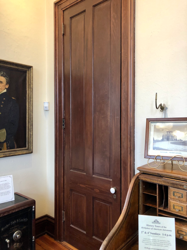 Ein Raum mit einer hölzernen Tür und einem Gemälde an der Wand, das einen Tisch mit einem Foto Rahmen und eine Tafel mit Text auf der linken Seite sowie Foto Rahmen und eine Schalttafel im Hintergrund zeigt, was auf einen Museums Eingang hindeutet.