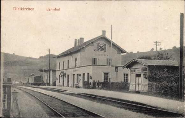 Schwarzes und weißes Foto eines Bahnhofs in Dielkirchen, Deutschland, mit einem Zug auf den Schienen, umgeben von Häusern, Pfählen, Bäumen, Pflanzen und Menschen, Hügel im Hintergrund und einem klaren Himmel.