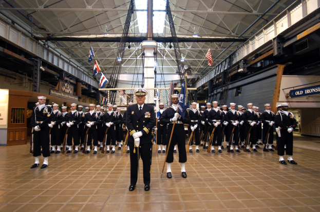 Eine Gruppe uniformierter Männer in einem großen Raum, die Mützen tragen und Gewehre halten, mit Flaggen und Schildern im Hintergrund.