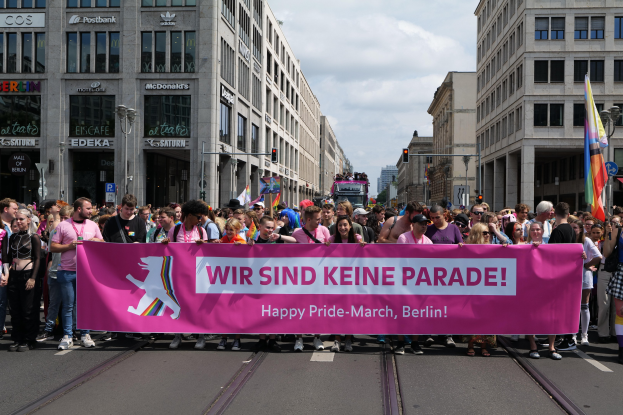 Eine Gruppe von Menschen marschiert auf einer Straße in Berlin mit einem pinken Banner, auf dem "Happy Pride March" steht, während Gebäude, Laternenmasten und Verkehrszeichen die Straße säumen und der Himmel bewölkt ist.