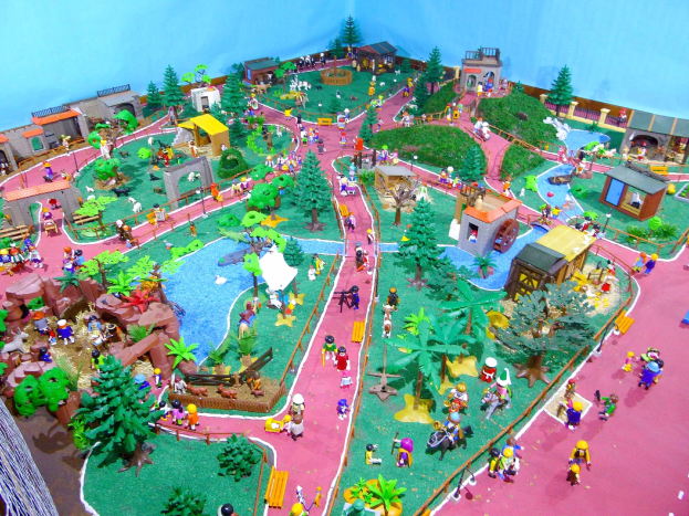 Zoo-Modell mit Miniatur-Leute, Bäumen, Häusern, Pflanzen, Gras, einem Zaun und einer Hintergrundwand, mit Text unten.
