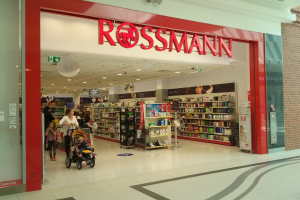 Ein Rossmann-Laden in einem Einkaufszentrum mit Menschen auf dem Boden, einer mit einem Kinderwagen, und Regalen mit Produkten unter Deckenleuchten und Schildern.