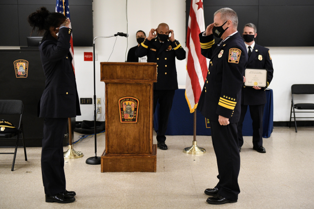 Gruppe uniformierter Personen bei einer Vereidigung eines neuen Feuerwehrchefs, der eine Urkunde hält, vor einem Mikrofon, Flaggen und Stühlen im Hintergrund.
