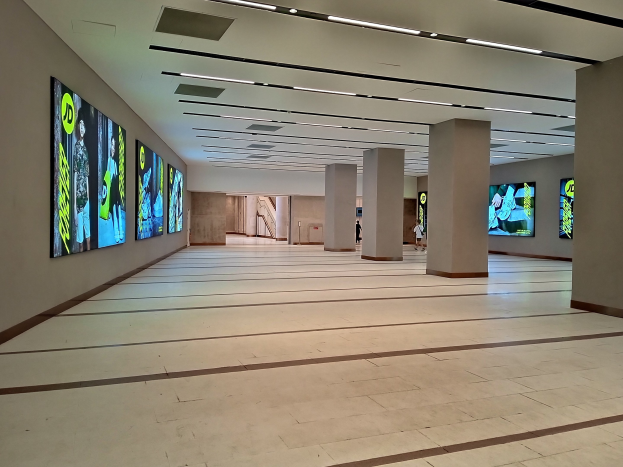 Ein großer Raum mit zahlreichen wandmontierten Fernsehern, die von Deckenlampen beleuchtet werden und ein paar Menschen sowie einen sichtbaren Boden aufweisen.