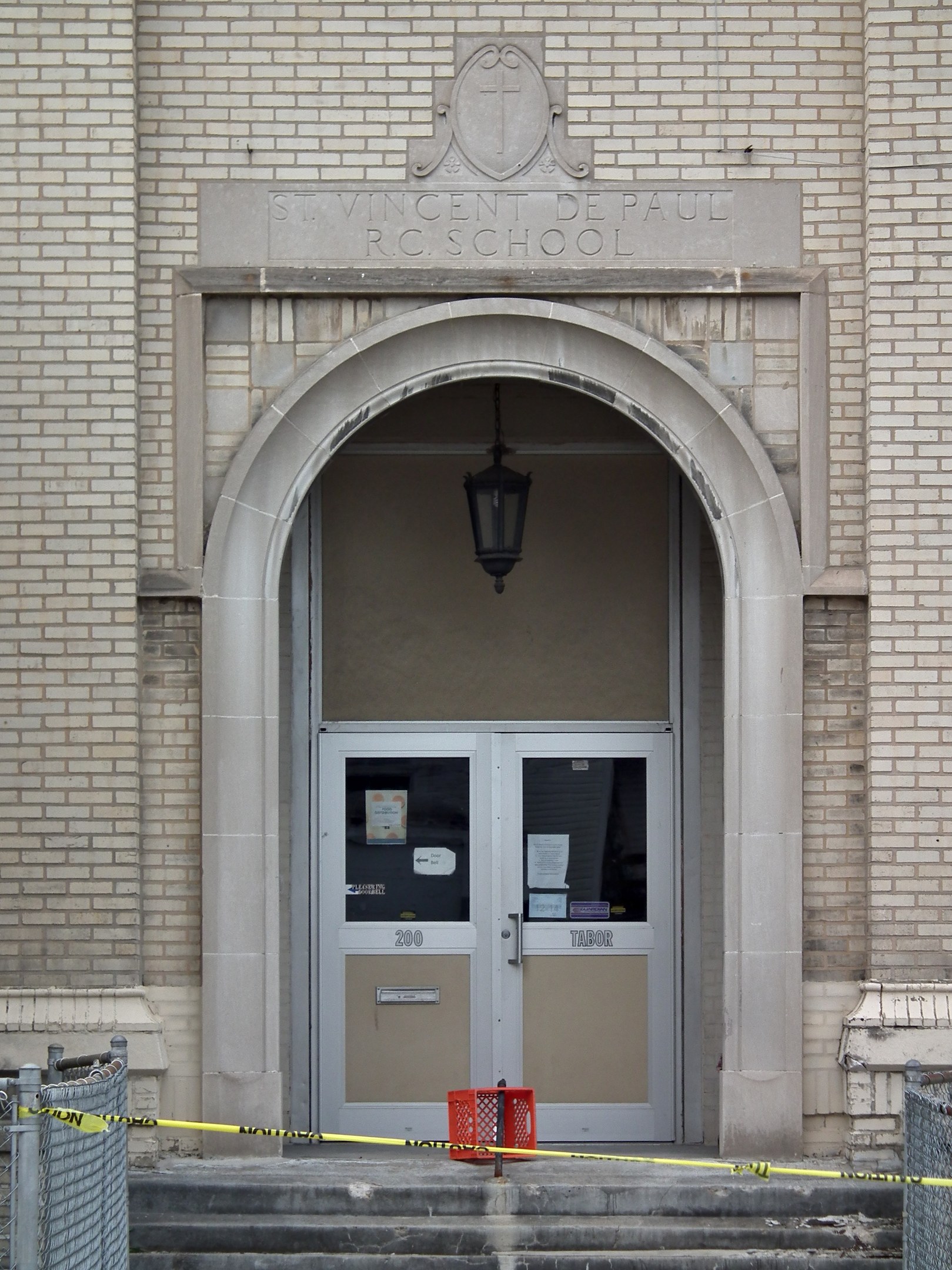 Eingang zu einem Backsteingebäude mit gelbem Absperrband, Treppe mit Maschendrahtzaun, einer Tür mit Papieren, einer hängenden Lampe und einem Gebäudenamen an der Wand.
