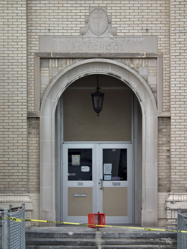 Eingang zu einem Backsteingebäude mit gelbem Absperrband, Treppe mit Maschendrahtzaun, einer Tür mit Papieren, einer hängenden Lampe und einem Gebäudenamen an der Wand.