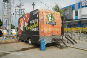 Lastwagen vor einem Gebäude mit einer Tafel, auf der "Teilen Sie eine positiv überladene Erfahrung" steht, mit Stufen, einem Zylinder auf dem Boden, Geländern, Strommasten mit Drähten, Laternen, Bäumen und einer bewölkten Himmel.