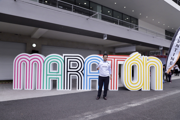 Ein Mann lächelt vor einem "Marathon"-Schild, umgeben von Menschen mit Schildern und einem beleuchteten Gebäude im Hintergrund.