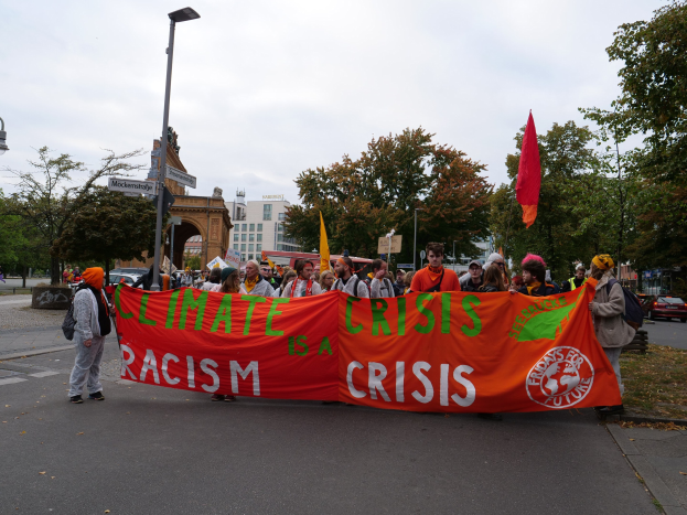Gruppe von Menschen marschiert auf einer von Bäumen gesäumten Straße und hält ein 'Klimakrise ist eine Krise'-Schild, mit Fahrzeugen, Gebäuden und einem klaren blauen Himmel im Hintergrund.