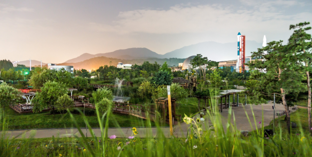 Ein Park mit grünem Gras, bunten Blumen, hohen Bäumen, einer Brücke mit Geländern, einem Springbrunnen, Laternenpfählen und Gebäuden im Hintergrund, vor Bergen und einem blauen Himmel.