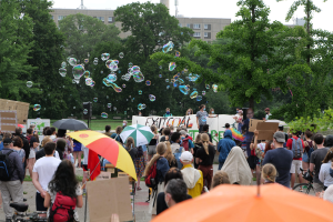 Eine Gruppe von Menschen, die vor einer Protestmenge mit Schirmen, Taschen und Schildern steht, mit Seifenblasen in der Luft, vor einer Kulisse aus Bäumen, Gebäuden, einem Turm und einem klaren blauen Himmel.