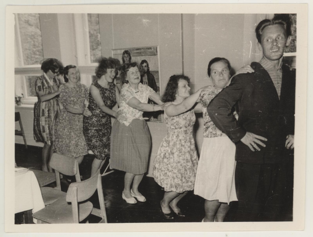 Eine Gruppe von Menschen, die in einem Raum mit Stühlen und einem Tisch tanzen, der mit einer weißen Decke bedeckt ist, sowie ein Foto auf dem Wand und Fenster im Hintergrund, gekleidet in 1950er-Jahre-Kleidung.