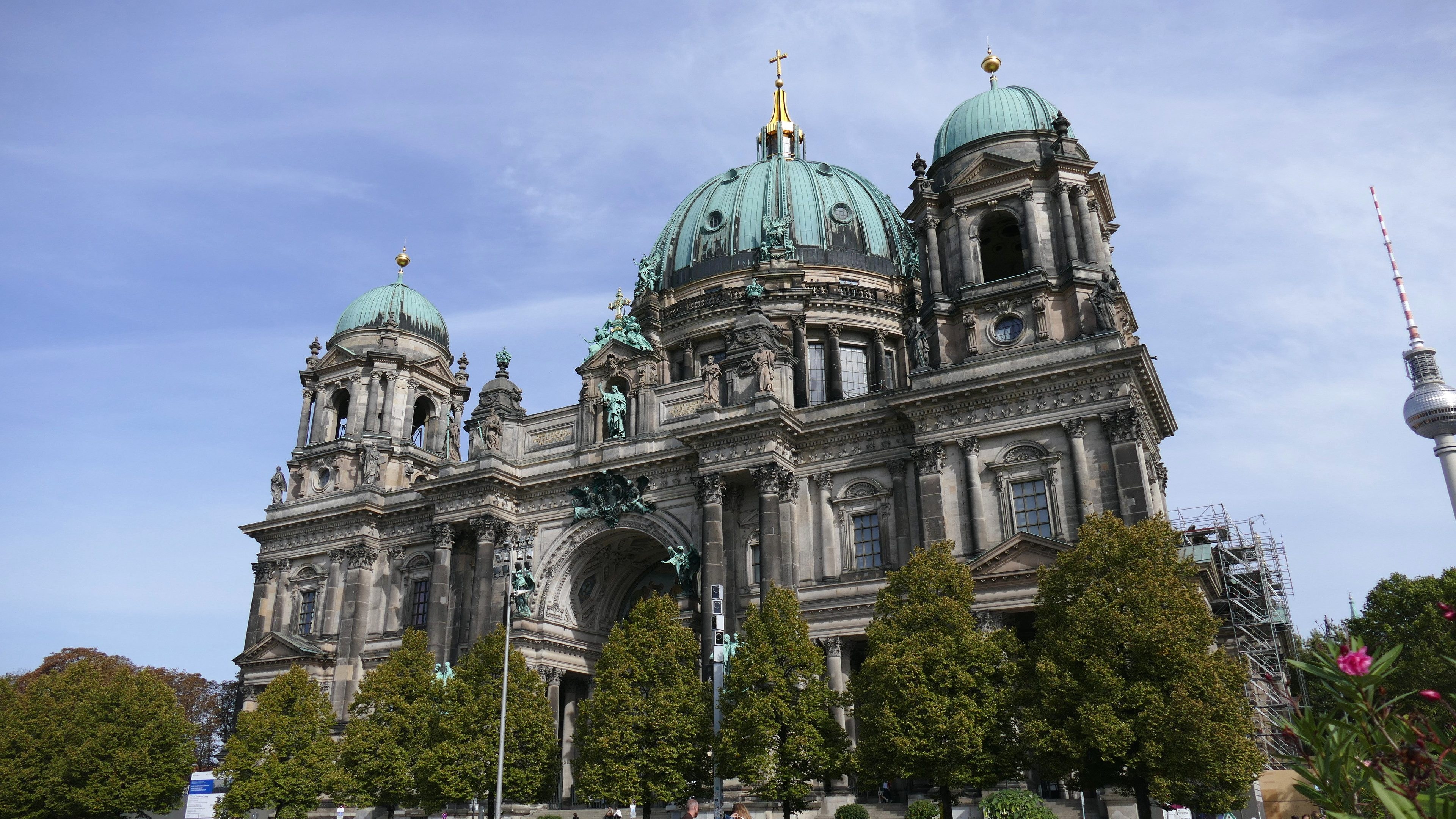 Großes, prunkvolles Berliner Dom-Gebäude in Berlin, Deutschland, mit architektonischen Details wie Fenstern, Säulen, Bögen und Statuen, vor einem bewölkten Himmel, mit Menschen, Bäumen und einem Turm im Vordergrund.
