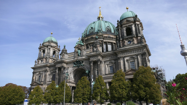 Großes, prunkvolles Berliner Dom-Gebäude in Berlin, Deutschland, mit architektonischen Details wie Fenstern, Säulen, Bögen und Statuen, vor einem bewölkten Himmel, mit Menschen, Bäumen und einem Turm im Vordergrund.