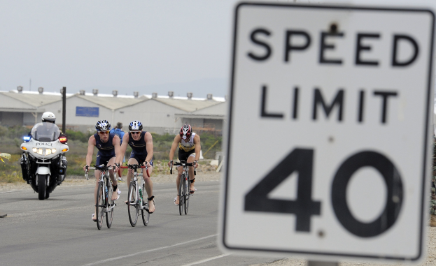 Eine Gruppe von Radfahrern mit Helmen fährt auf einer Straße neben einem Geschwindigkeitsbegrenzungsschild vorbei, mit Gebäuden, Strommästen, Vegetation und einem klaren blauen Himmel im Hintergrund.