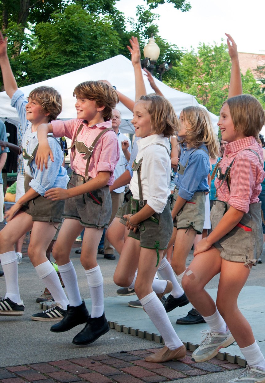 Kinder in traditioneller bayrischer Tracht tanzen auf einer Straße, umgeben von einer Menge, Zelten und Gebäuden unter einem klaren blauen Himmel.