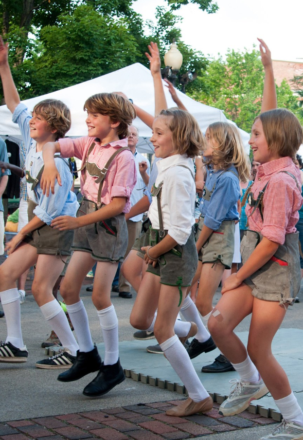 Kinder in traditioneller bayrischer Tracht tanzen auf einer Straße, umgeben von einer Menge, Zelten und Gebäuden unter einem klaren blauen Himmel.