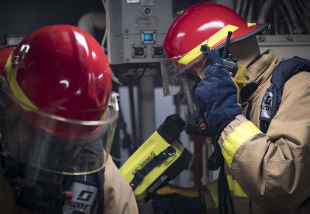 Zwei Feuerwehrleute in Schutzausrüstung bei der Arbeit an einem Hydranten während einer Übung mit Maschinen und Kabeln im Hintergrund.