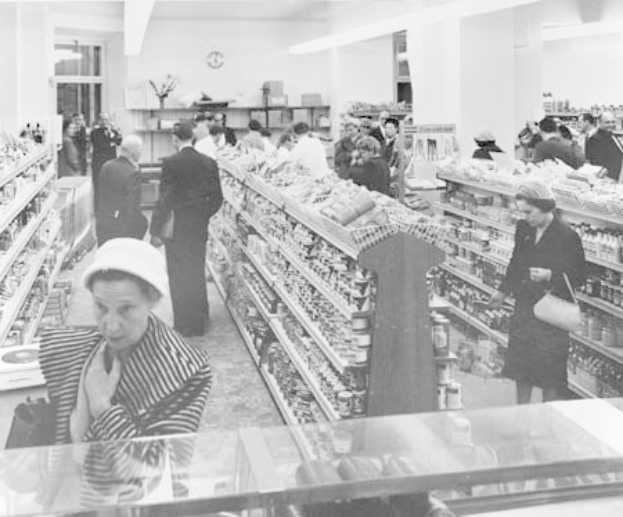 Schwarzes und weißes Foto von Menschen beim Einkaufen in einem Supermarkt, die Taschen halten, mit Lebensmittelregalen, einer Wanduhr und Deckenlampen.