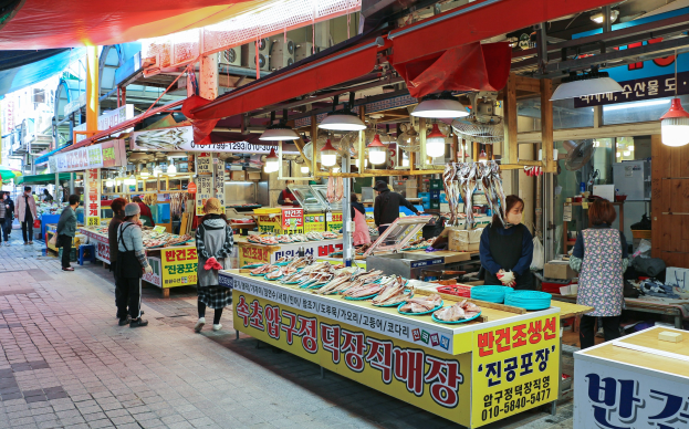 Ein belebter Straßenmarkt in Seoul, Südkorea, mit Menschen, Tischen mit verschiedenen Gegenständen und Gebäuden im Hintergrund bei klarem blauem Himmel.