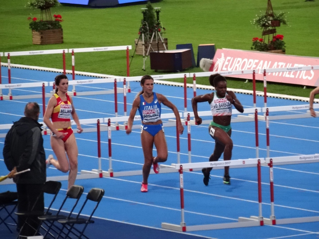 Gruppe von Frauen, die Hürden auf einem Rasenlaufbahn laufen, mit einer Person auf der linken Seite, die einen Stab hält, Stühlen und einem Schreibtisch im Vordergrund, Blumentöpfe und Texttafeln im Hintergrund.