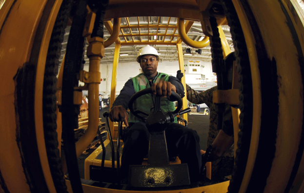 Ein Mann in einer grünen Jacke und einem weißen Helm bedient einen Gabelstapler in einer Fabrik, mit anderen Menschen im Hintergrund.