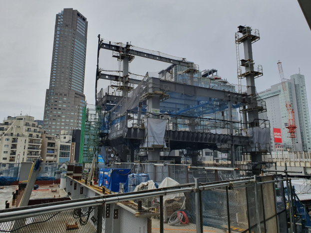 Großes Gebäude im Bau auf einem städtischen Kraftwerksgelände, umgeben von einem Metallzaun, benachbarten Gebäuden, Versorgungsmasten, einer Tafel, Fahrzeugen, Überlandleitungen und einem bewölkten Himmel.