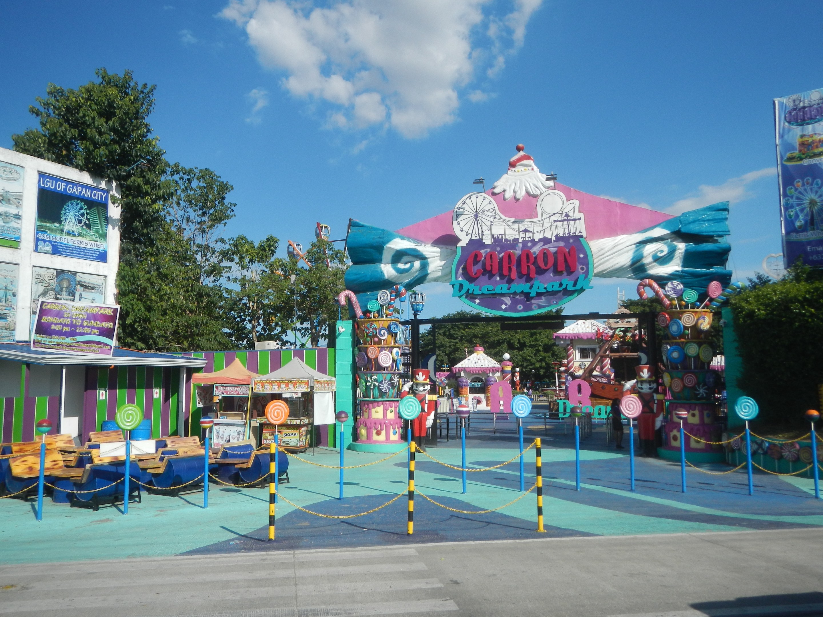 Karussell am Parkeingang mit Straße, Umzäunung, Buden, Banner, Bäumen und bewölktem Himmel.
