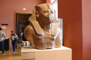 Eine Büste eines ägyptischen Pharaos in einem Museum ausgestellt, umgeben von Menschen, einer Texttafel, einer Tür und Deckenleuchten.