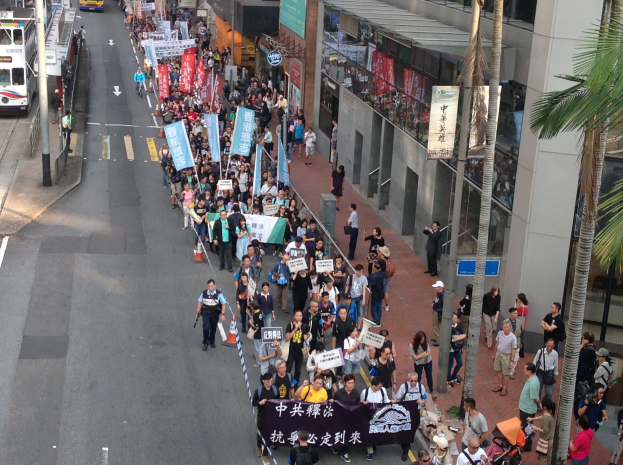 Eine große Gruppe von Menschen marschiert auf einer Stadtstraße mit Schildern und Transparenten, mit Bäumen, Gebäuden und Fahrzeugen auf beiden Seiten.