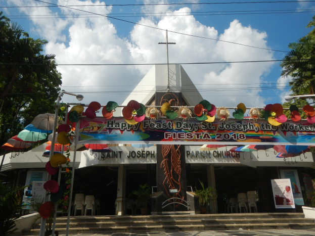 Gebäude mit einem "Frohen Festtag St. Josef der Arbeiter"-Schild, Treppe zum Eingang, ein Bogen mit hängenden Dekorationen, Außenbereich mit Tischen und Stühlen, Topfpflanzen, Bäume auf beiden Seiten und ein bewölkter Himmel.