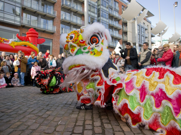 Ein lebendiges chinesisches Neujahrsfest in Amsterdam mit einem Löwendance im Vordergrund und einer Menschenmenge, einige halten Kameras, vor einem Hintergrund aus Gebäuden, Laternenmästen und einem klaren blauen Himmel.