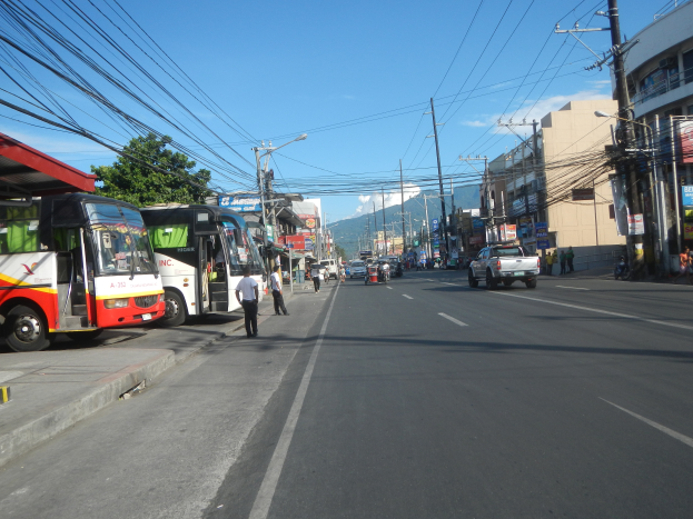 Eine belebte Stadtstraße mit zahlreichen Fahrzeugen, Fußgängern auf den Gehwegen, Gebäuden, Bäumen, Strommasten und fernen Hügeln unter einem bewölkten Himmel.