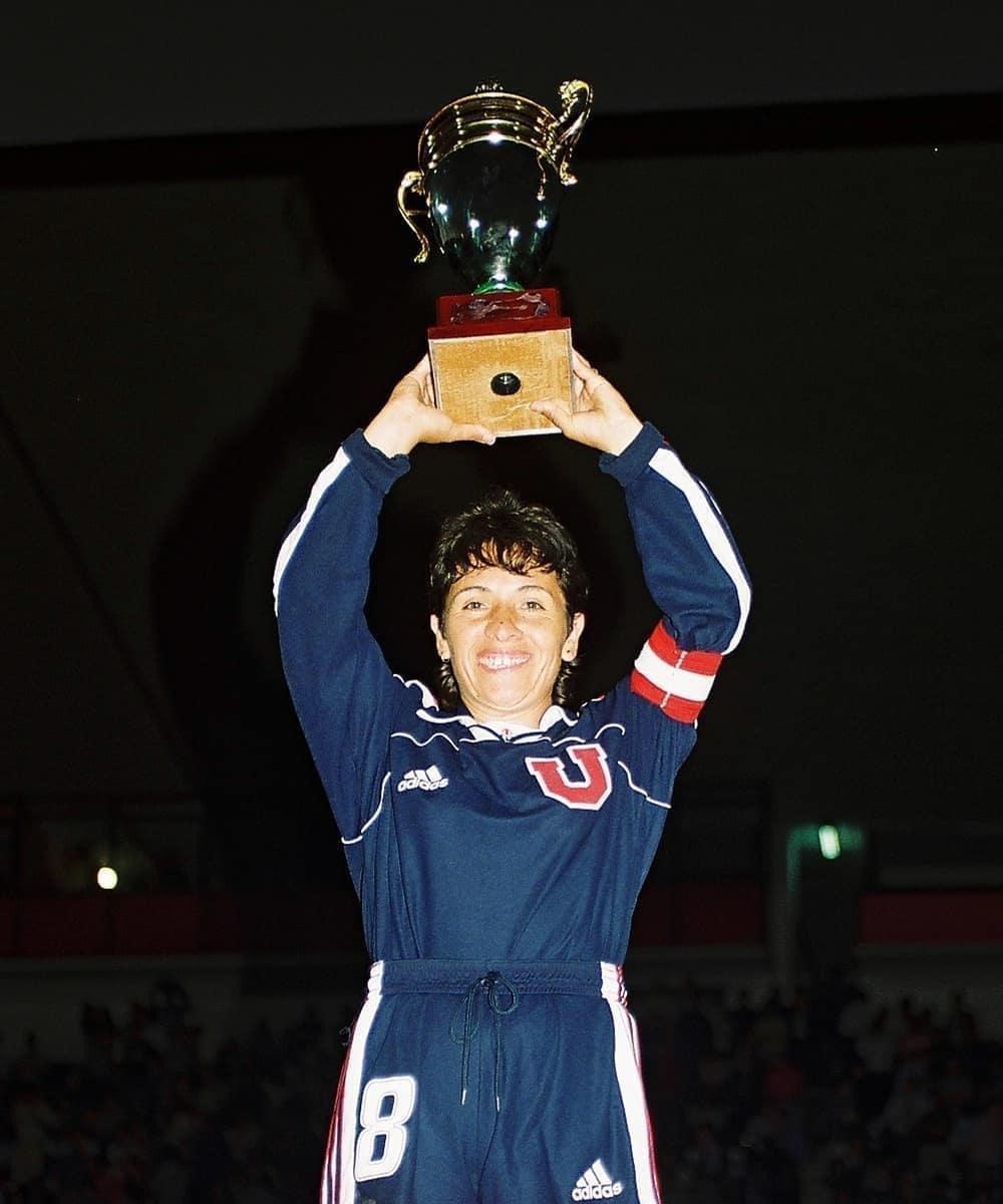 Eine Frau in einem blauen Kleid hält eine Trophäe hoch und lächelt, umgeben von einer jubelnden Menge in einem hell erleuchteten Stadion.