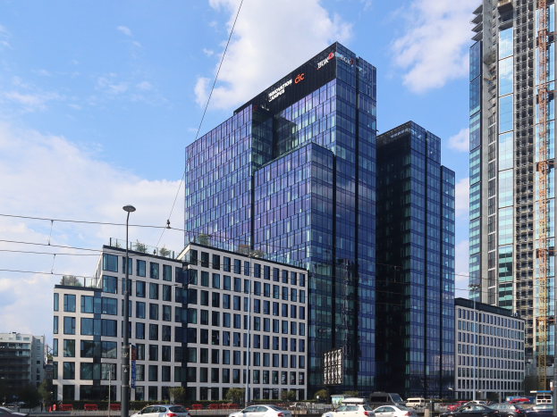 Großes modernes Bürogebäude mit zahlreichen Fenstern, identifiziert als Deutsche Bank Hauptsitz in Frankfurt, Deutschland, in einer städtischen Umgebung mit Bäumen, Straßeninfrastruktur, Fahrzeugen und bewölktem Himmel.