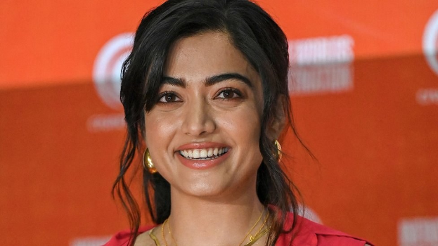 Eine Frau in einem leuchtend roten Kleid mit einem eleganten Dutt, schwarzen High Heels und dezentem Make-up lächelt in die Kamera und steht vor einem hellgelben Banner mit der Aufschrift "Sonam Kapoors beste Schönheits Looks vom Cannes Film Festival."