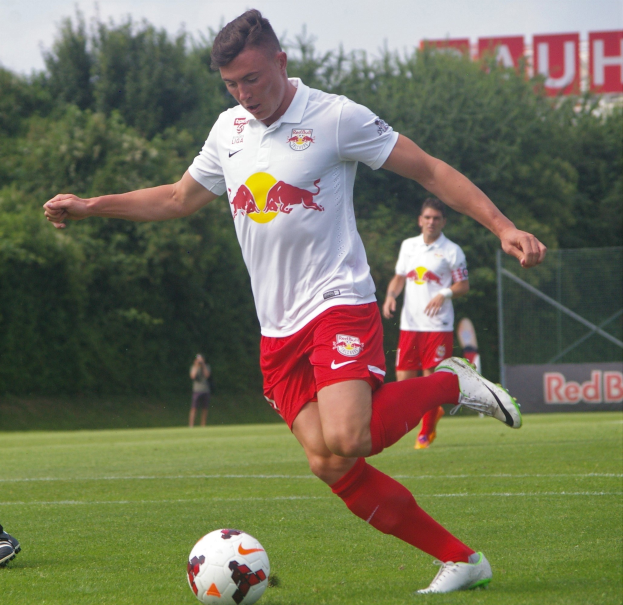 Ein professioneller Fußballspieler in einem roten Trikot tritt einen Fußball auf einem grünen Feld mit Bäumen und einem klaren blauen Himmel im Hintergrund, wobei ein Schild mit der Aufschrift "RB Leipzig" im Hintergrund zu sehen ist.