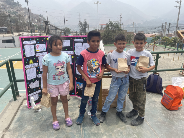 Gruppe von Kindern auf einem Basketballfeld mit Papiertüten in den H├Ąnden, mit Gegenst├Ąnden auf der rechten Seite und einer Tafel mit Text auf der linken Seite, vor einer ├Ąu├čeren Kulisse mit Zaun, Pfosten und Geb├Ąuden.