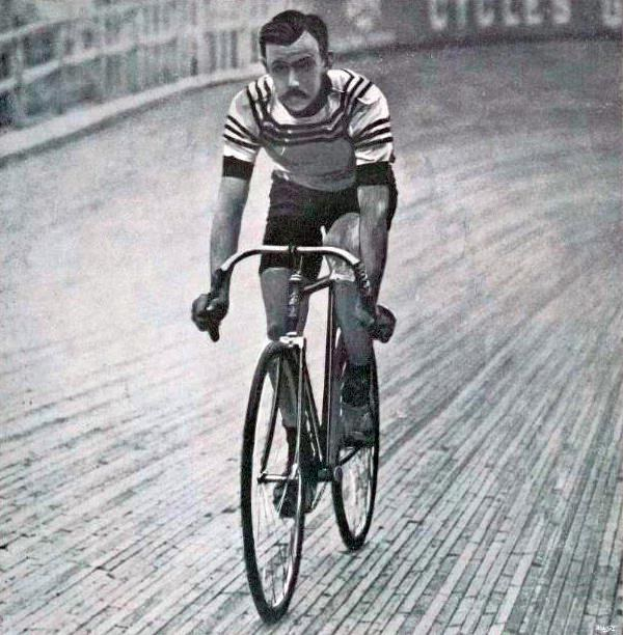 Schwarz-weiß-Foto eines Mannes, der auf einer Bahn mit einem Zaun und einer Tafel im Hintergrund Fahrrad fährt.
