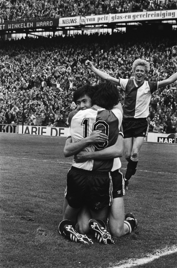 Drei Fußballspieler umarmen sich auf dem Boden nach einem Tor, mit jubelnder Menge und schwenkenden Fahnen im Hintergrund, in einer schwarz-weißen Stadionatmosphäre.