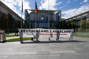 Gruppe von Menschen mit einem Banner mit der Aufschrift "Deutschland, Lagerland Migration ist kein Verbrechen" vor einem Gebäude mit Fenstern, umgeben von Gras, einem Metallzaun, Pflanzen, Bäumen und Fahnenstangen unter einem bewölkten Himmel.