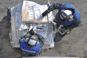 Zwei Männer in blauen Uniformen und Helmen arbeiten an einem großen Drohnenmodell, hältend verschiedene Gegenstände, während eine Plastikabdeckung auf dem Boden liegt.