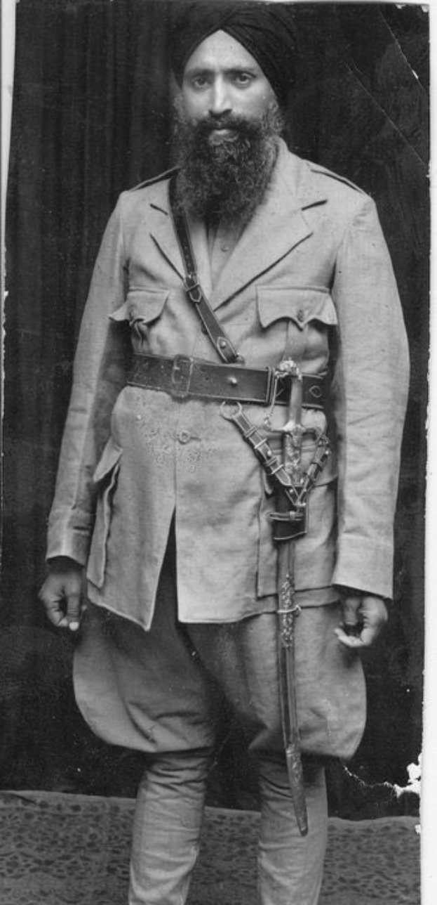 Schwarzes und weißes Porträt eines Mannes mit langem Bart, Turban und Schwert, der als Guru Nanak Dev Ji identifiziert wird, vor einem Vorhang steht.