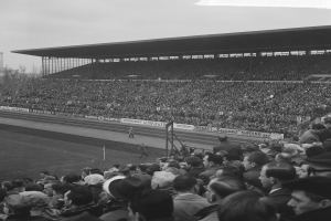 Schwarzes und weißes Foto eines vollen Stadions mit Zuschauern, die ein Fußballspiel beobachten, einige stehen und andere sitzen auf den Tribünen, mit Bannern, Pfählen, einer Hütte, Bäumen, einem Turm und einem bewölkten Himmel im Hintergrund.